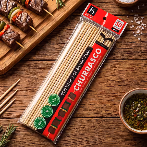 Pacote de espetinho de bambu para churrasco Kozin com 50 unidades sobre mesa de madeira, ao lado de espetos de carne e legumes