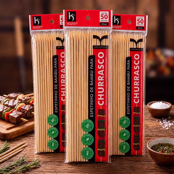 Três pacotes de espetinho de bambu para churrasco Kozin com 50 unidades de 25 cm, sobre mesa de madeira com espetos de carne